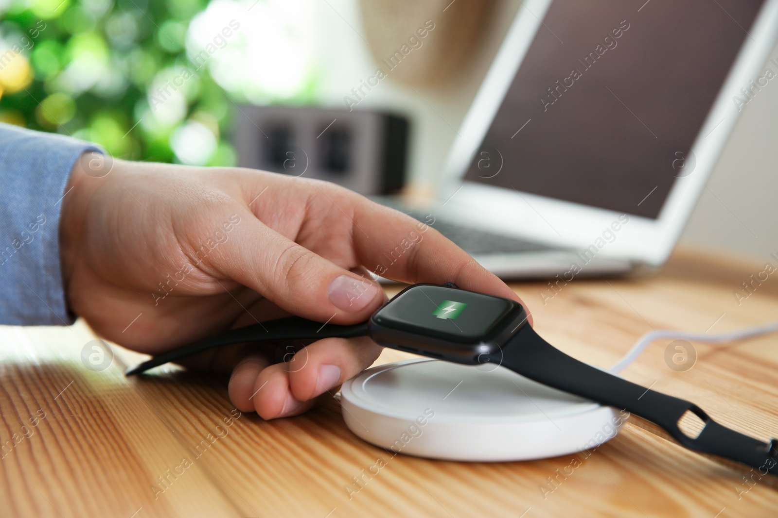 Photo of Man putting smartwatch onto wireless charger at wooden table, closeup. Modern workplace accessory
