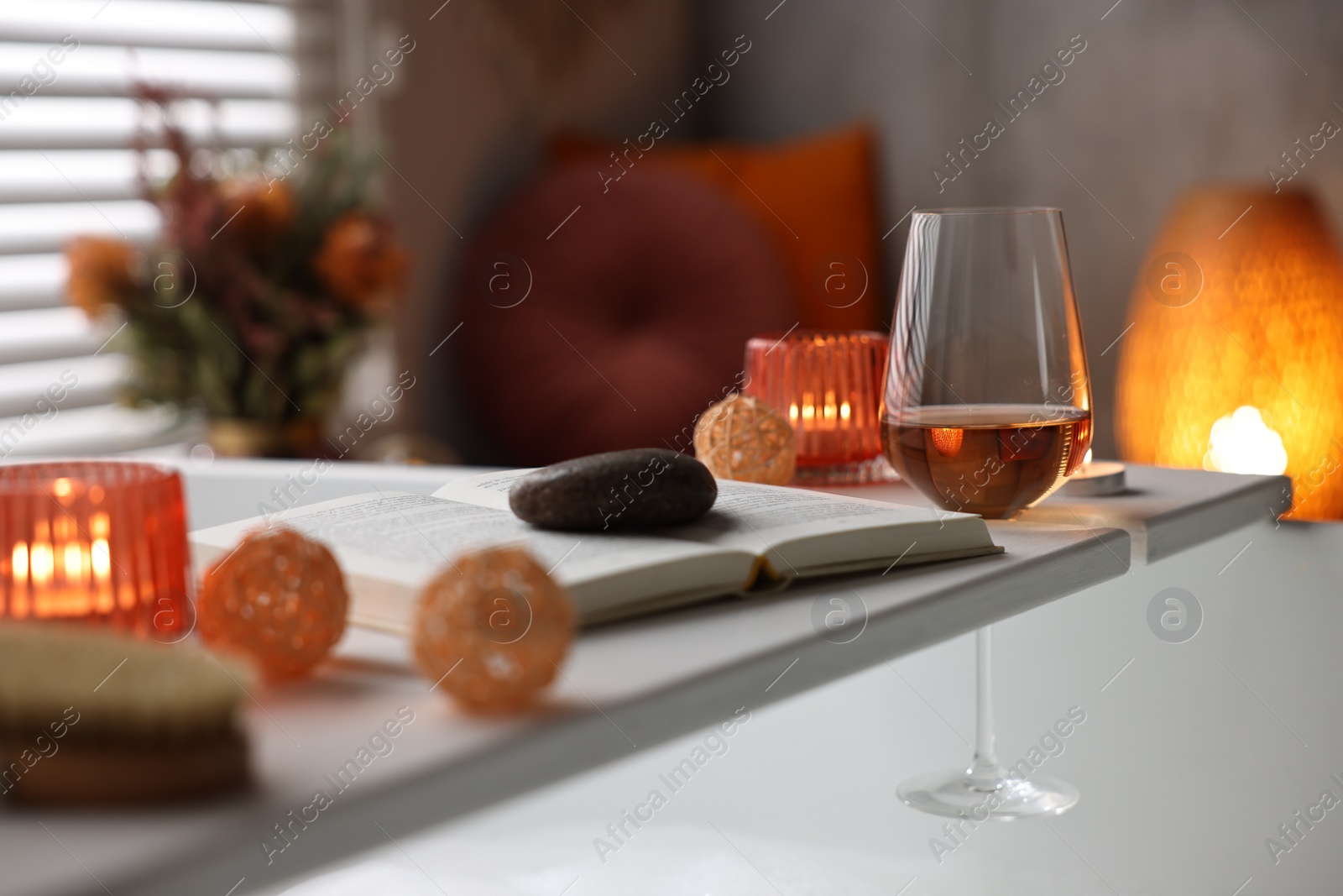 Photo of White wooden tray with glass of rose wine, book and burning candles on bathtub in bathroom