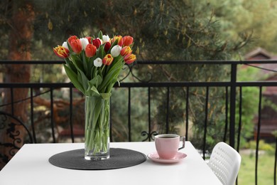 Photo of Beautiful colorful tulips in glass vase and cup of drink on white table at balcony, space for text