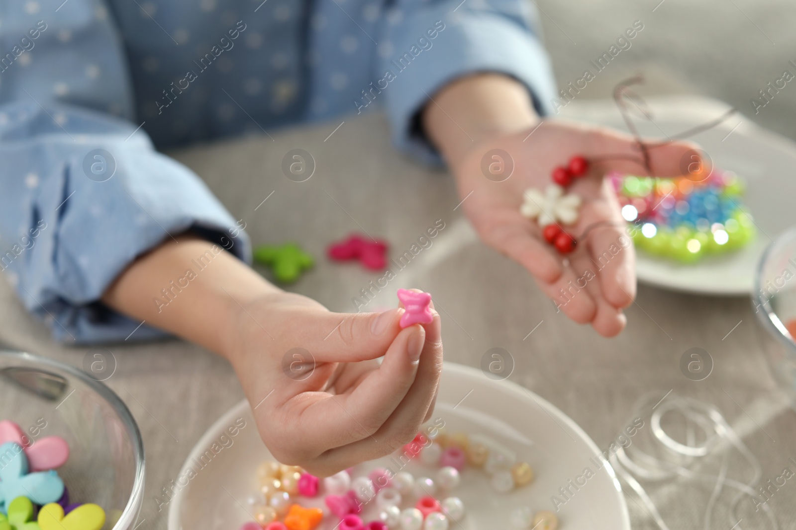 Photo of Little girl making accessory with beads at table, closeup. Creative hobby
