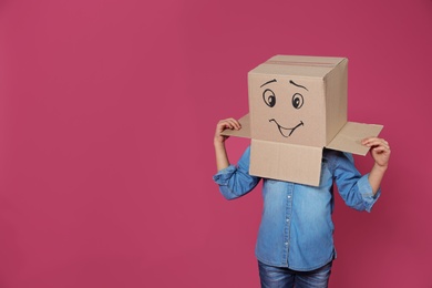 Cute little child wearing cardboard box with smiling face on color background. Space for text