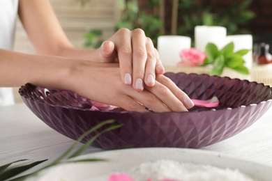 Photo of Woman soaking her hands in bowl with water and petals on table, closeup. Spa treatment