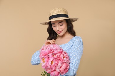 Beautiful young woman in straw hat with bouquet of pink peonies against beige background