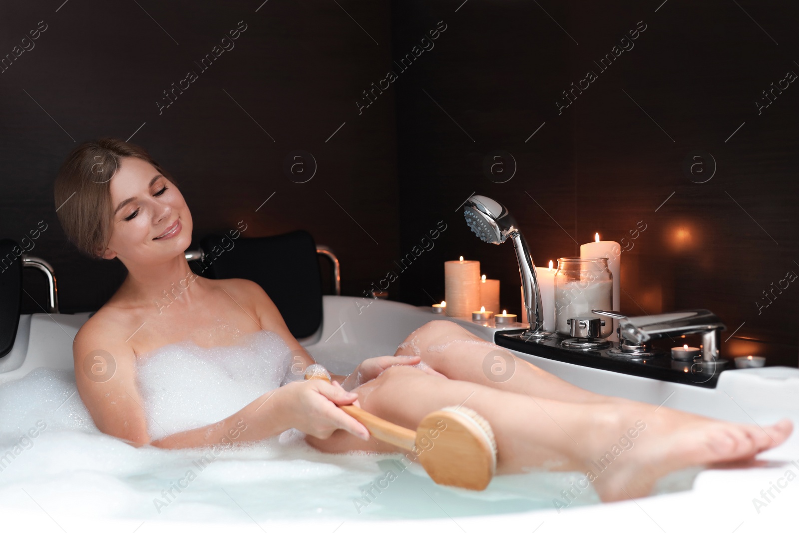 Photo of Beautiful woman rubbing leg with brush while taking bubble bath. Romantic atmosphere