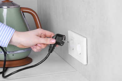 Photo of Woman plugging electric kettle into power socket at white table indoors, closeup