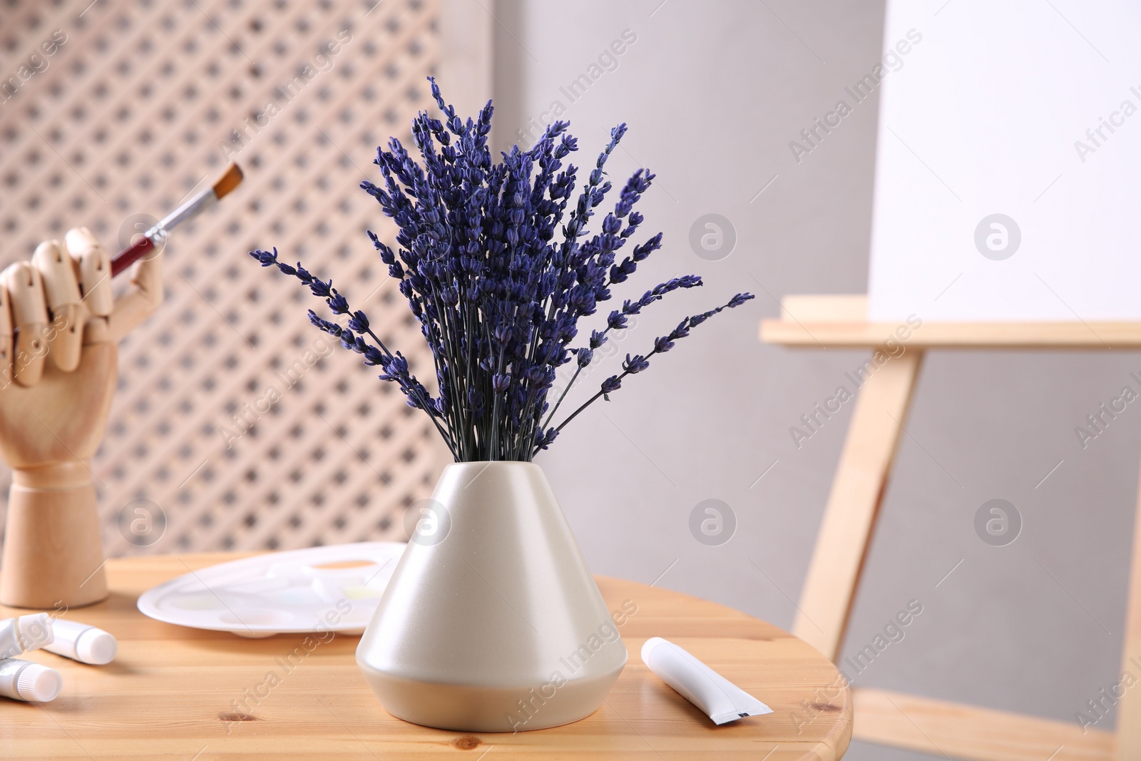 Photo of Bouquet of beautiful preserved lavender flowers, palette, paints and manikin hand with brush on wooden table near easel indoors, closeup