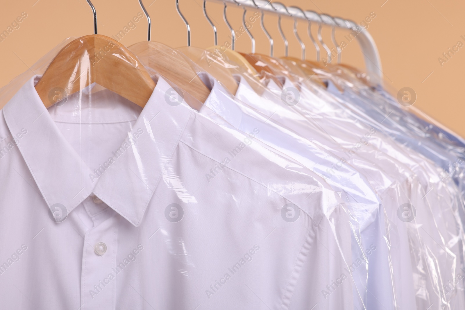 Photo of Dry-cleaning service. Many different clothes in plastic bags hanging on rack against beige background, closeup