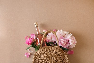 Photo of Wicker bag with bottle of rose wine and beautiful pink peonies on brown background, top view. Space for text