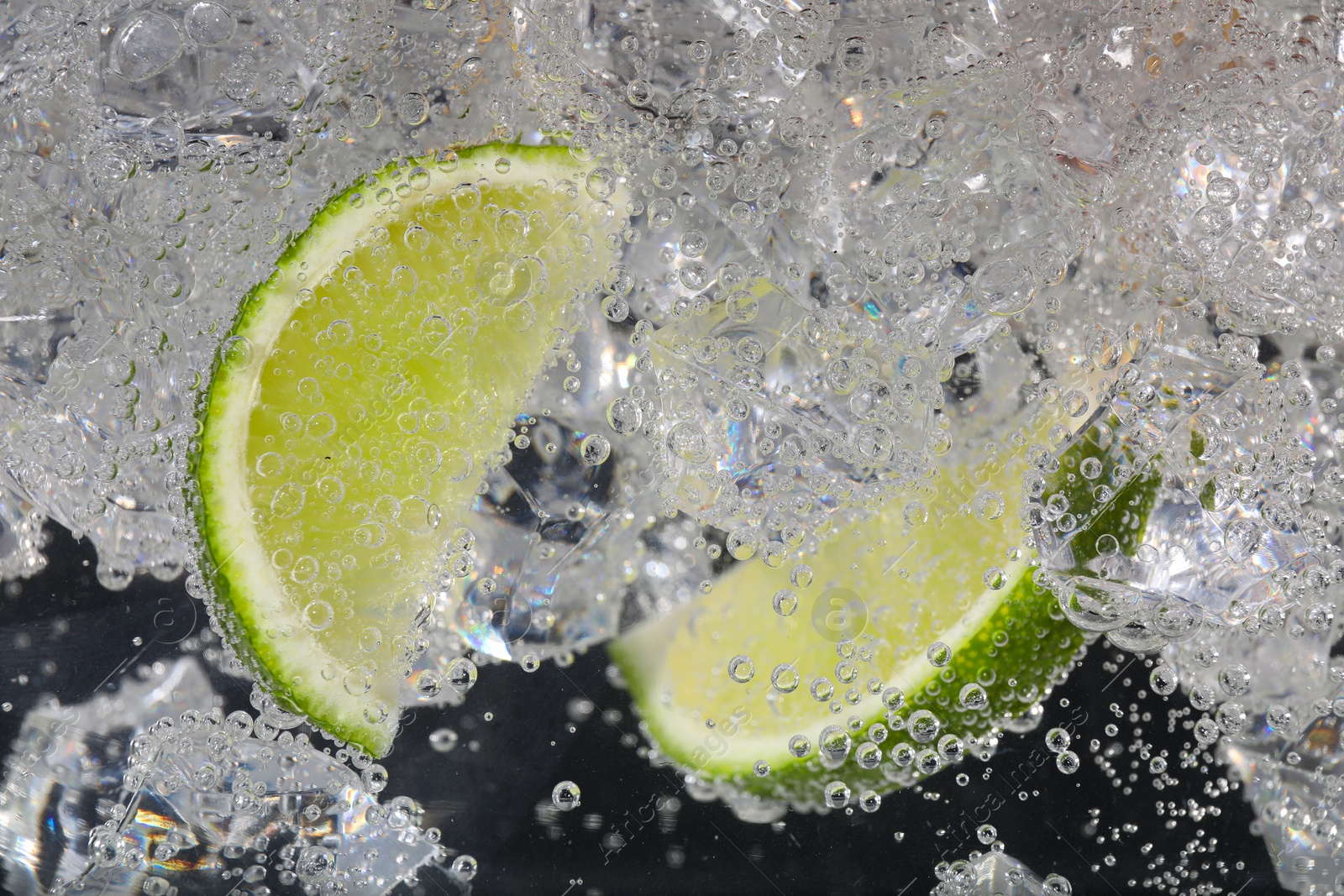 Photo of Juicy lime slices and ice cubes in soda water against black background, closeup