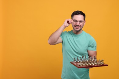 Smiling man holding chessboard with game pieces on orange background, space for text
