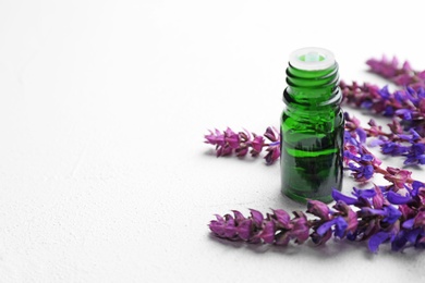 Photo of Bottle of sage essential oil and flowers on light table, space for text