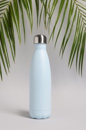 Stylish thermo bottle near tropical leaves on light grey background