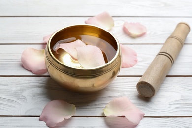Photo of Golden singing bowl with petals and mallet on white wooden table. Sound healing