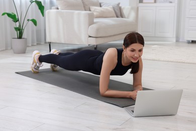 Photo of Happy woman doing plank exercise and watching video tutorial via laptop at home