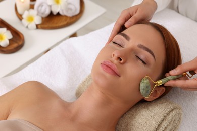 Photo of Young woman receiving facial massage with jade roller in beauty salon, closeup