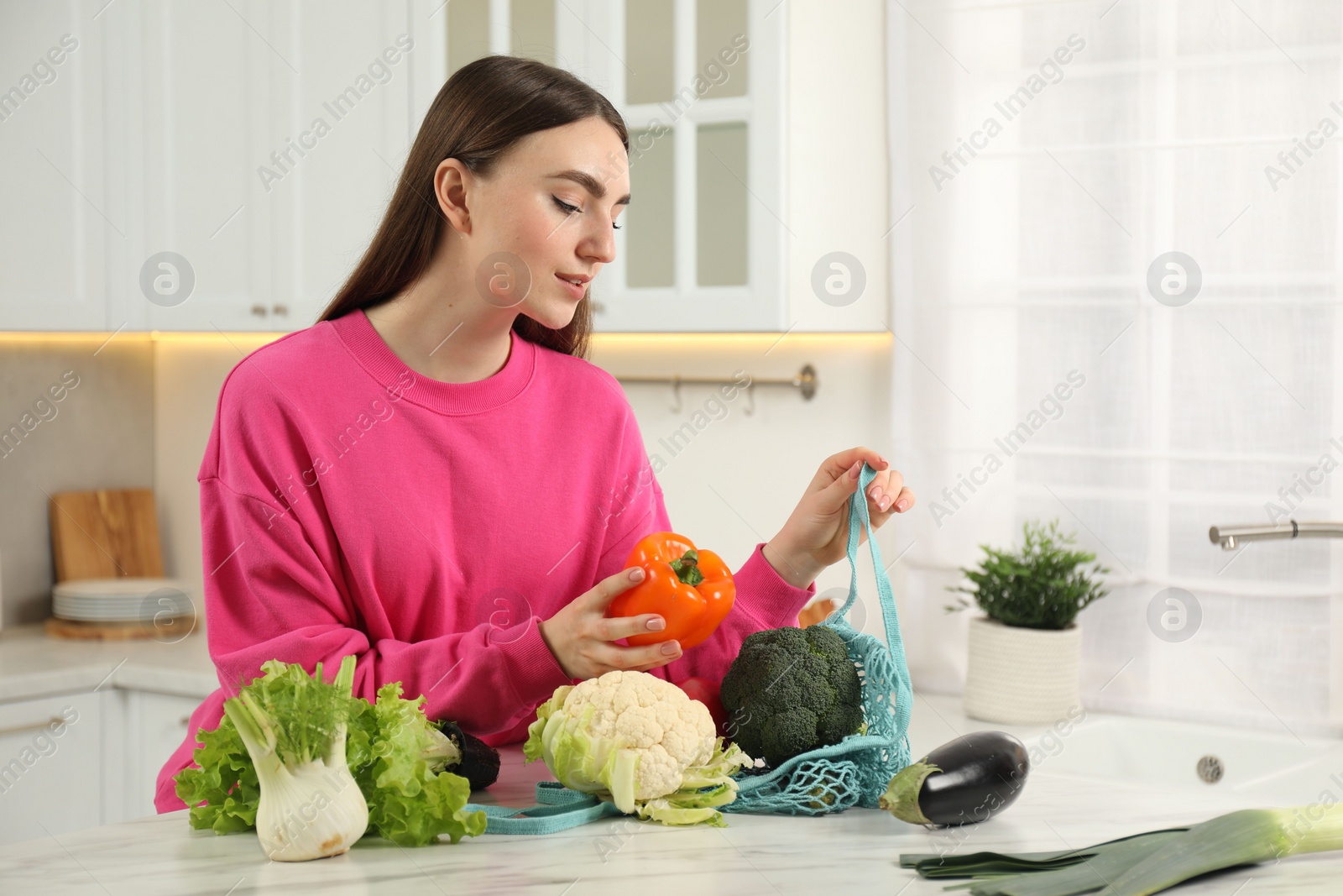 Photo of Woman taking pepper out from string bag at light marble table in kitchen