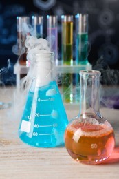 Photo of Laboratory glassware with colorful liquids and steam on white wooden table against black background, closeup. Chemical reaction