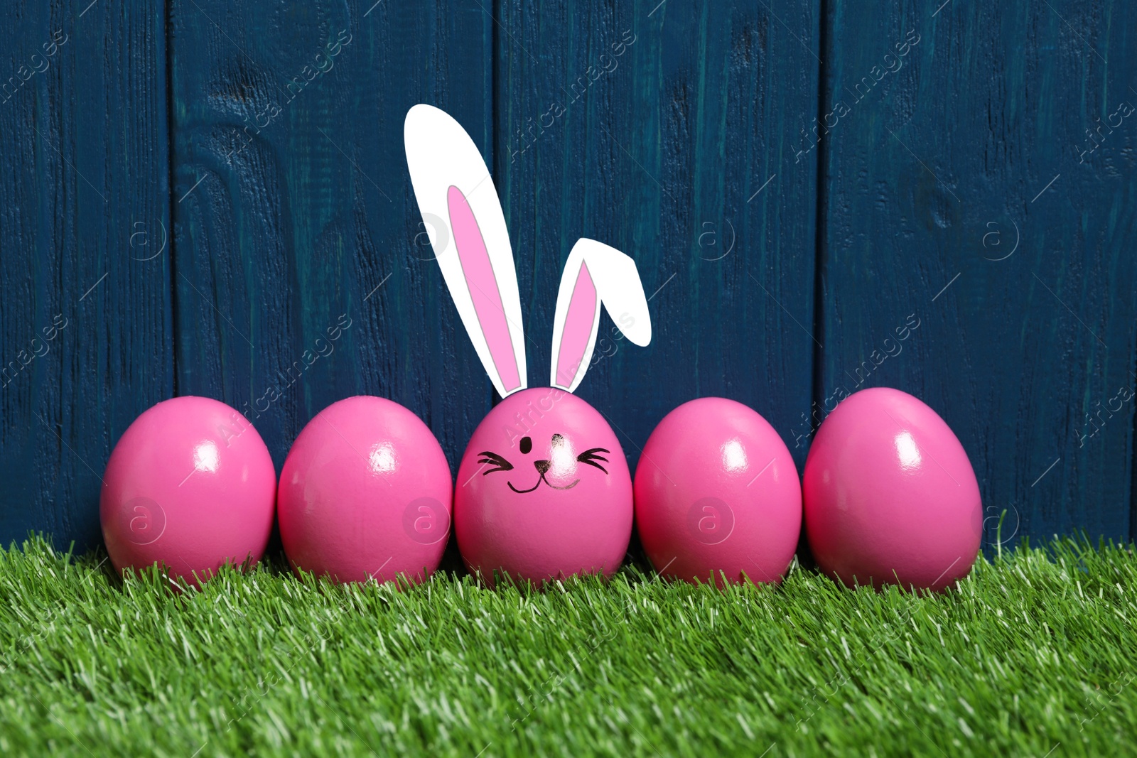 Image of One egg with drawn face and ears as Easter bunny among others on green grass against blue wooden background