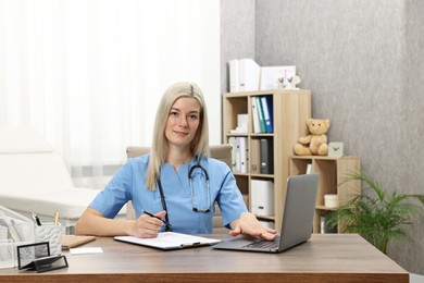 Photo of Doctor working with clipboard and laptop at table in clinic