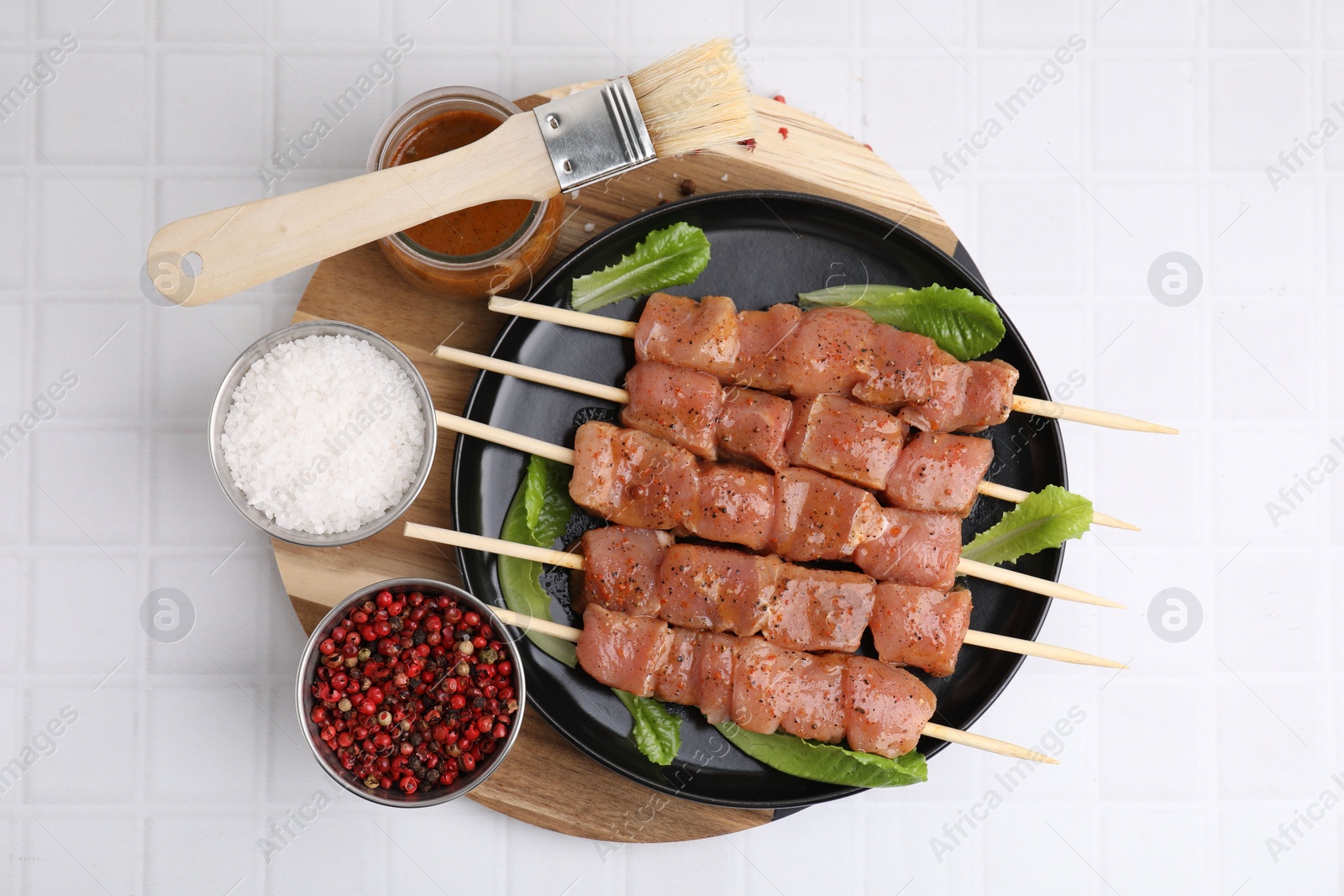Photo of Wooden skewers with cut raw marinated meat and basting brush on white tiled table, flat lay