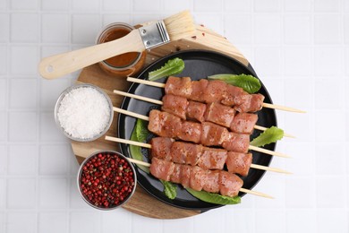 Wooden skewers with cut raw marinated meat and basting brush on white tiled table, flat lay