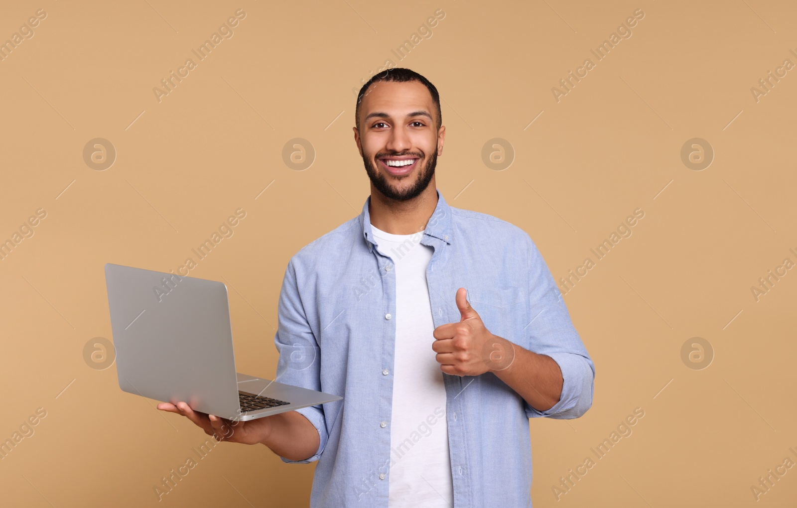 Photo of Smiling young man with laptop showing thumbs up on beige background