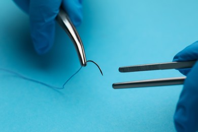 Professional surgeon holding forceps with suture thread on light blue background, closeup. Medical equipment