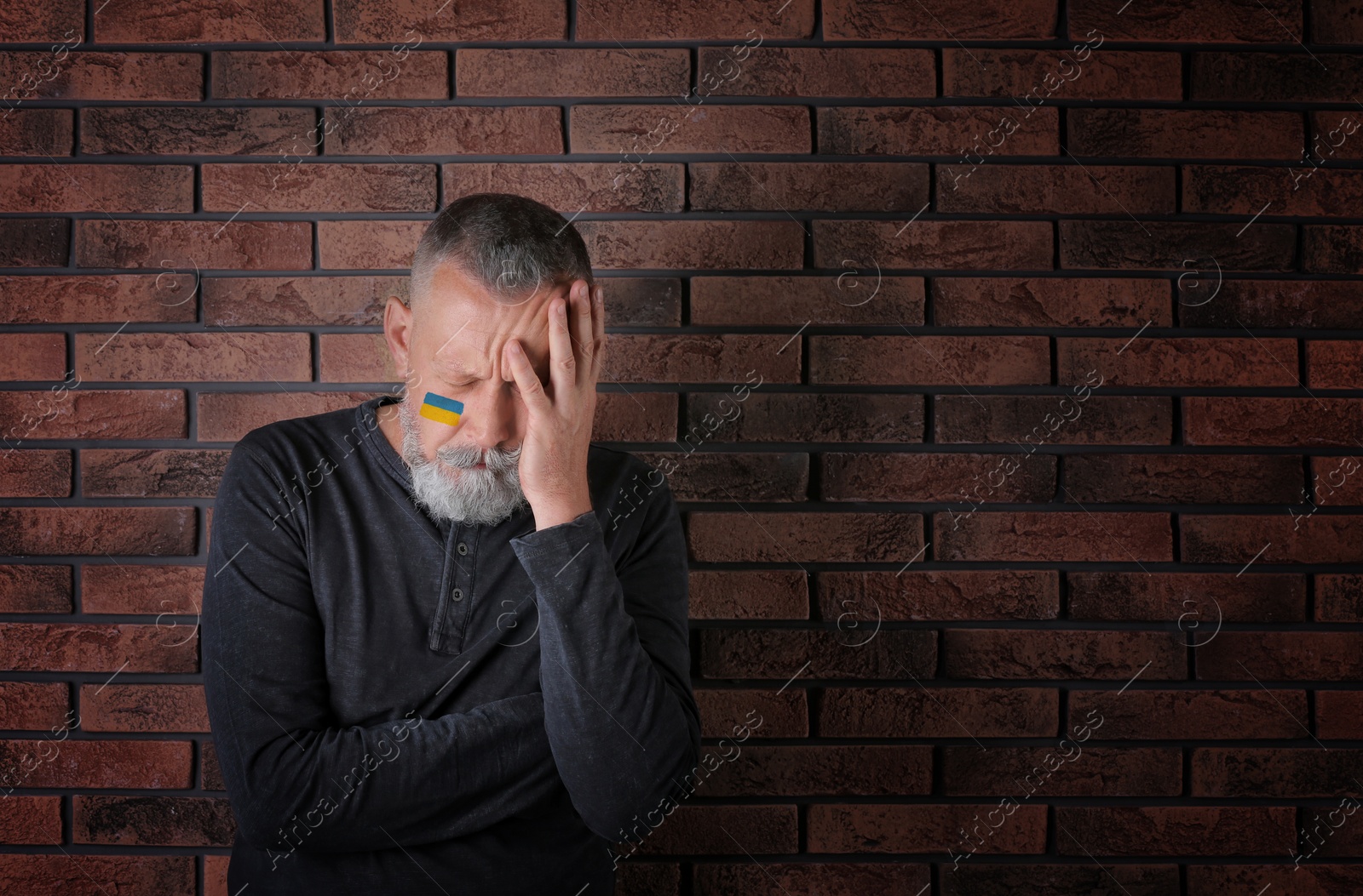 Image of Sad senior man with picture of Ukrainian flag on cheek near brick wall, space for text. Stop war in Ukraine