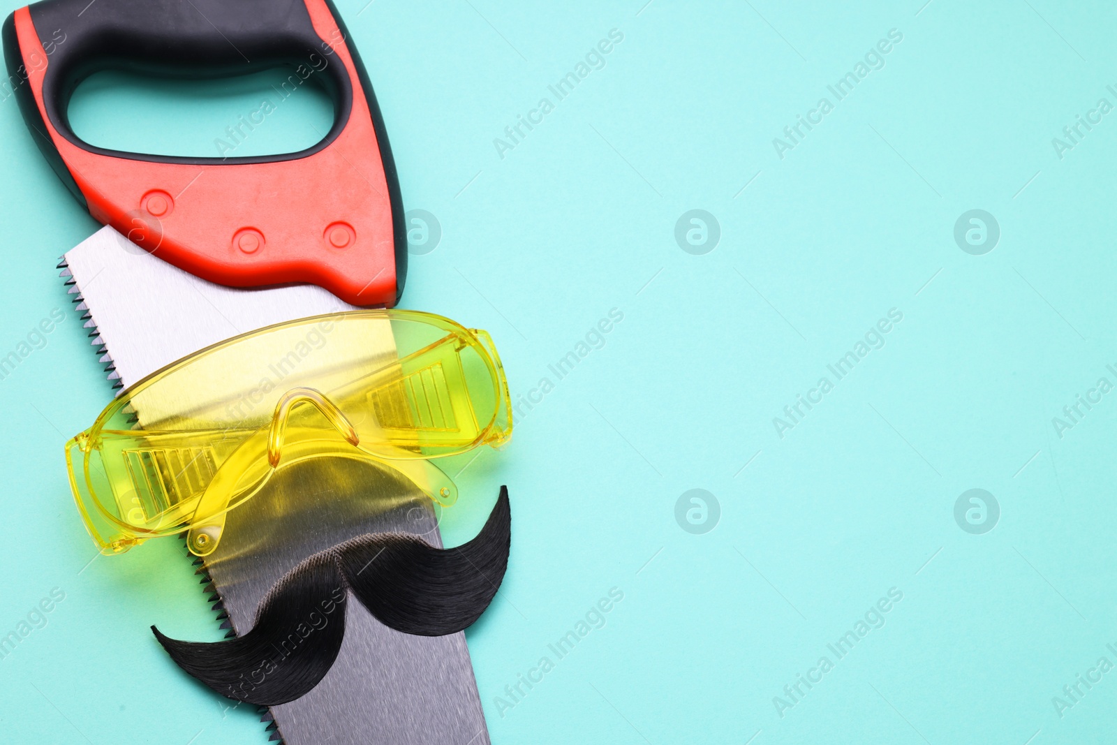 Photo of Man's face made of artificial mustache, safety glasses and hand saw on light blue background, top view. Space for text