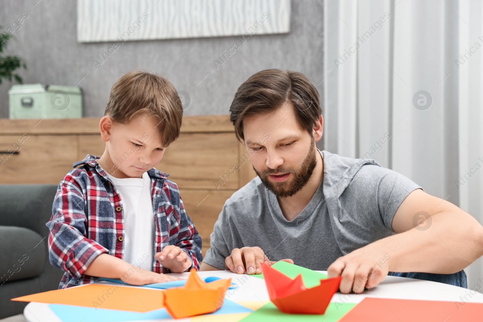 Photo of Dad and son making paper boats at coffee table indoors