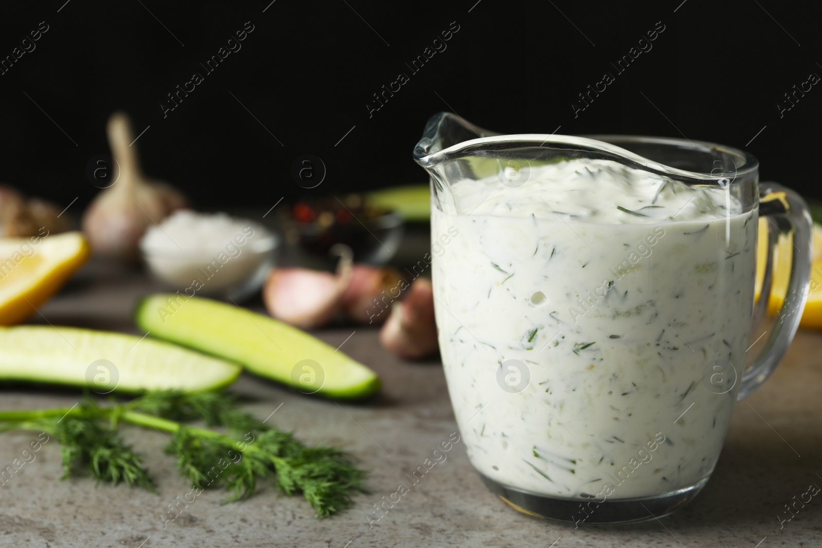 Photo of Glass jug of Tzatziki cucumber sauce with ingredients on table. Space for text