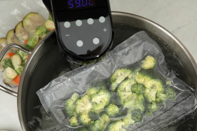 Photo of Sous vide cooker and vacuum packed broccoli in pot on white table, closeup. Thermal immersion circulator