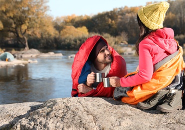 Photo of Couple of campers in sleeping bags sitting on rock near pond. Space for text