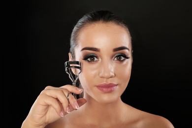 Portrait of young woman with eyelash extensions holding curler on black background