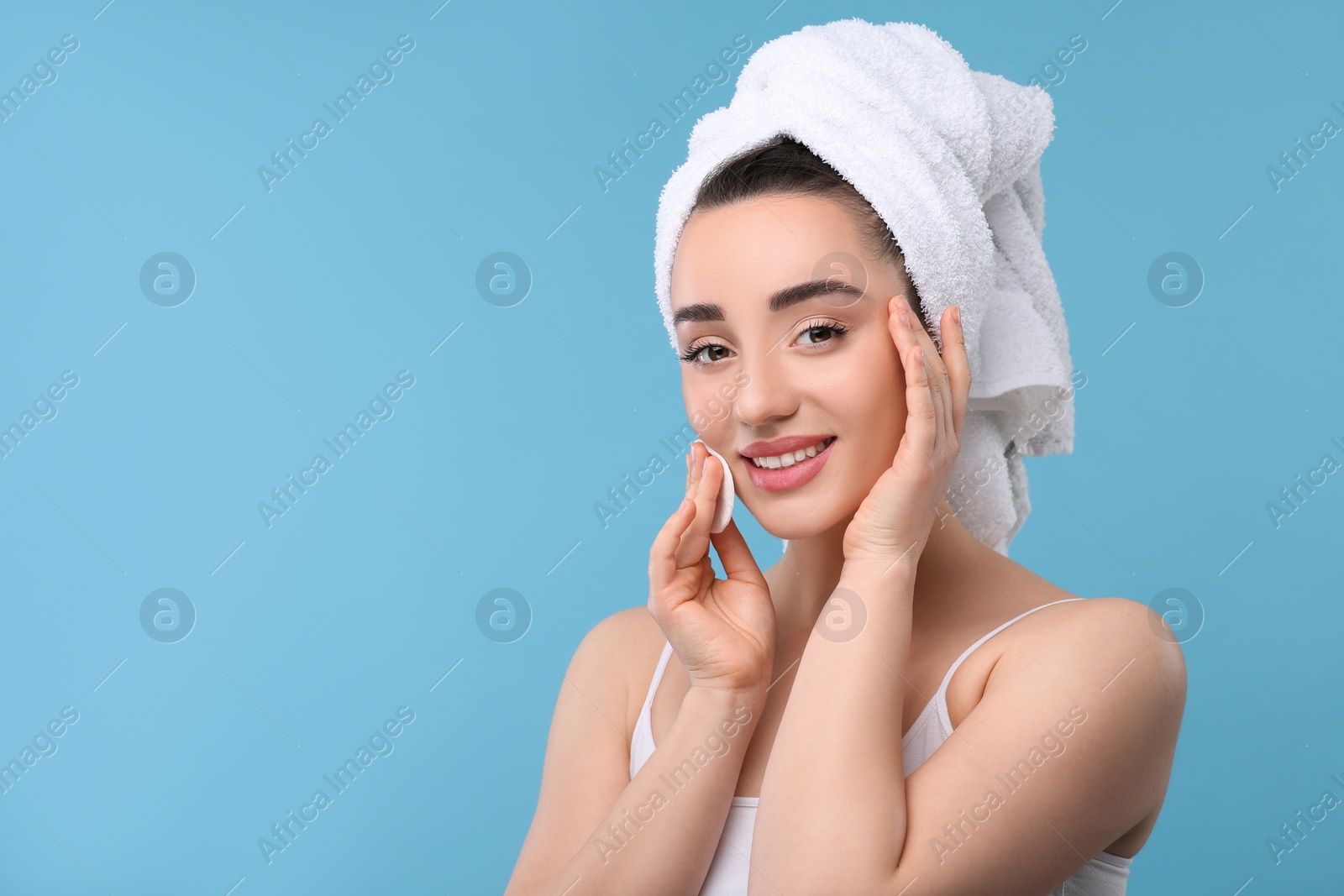 Photo of Beautiful woman in terry towel removing makeup with cotton pad on light blue background, space for text