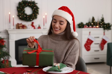 Photo of Happy young woman in Santa hat opening Christmas gift at served table in decorated room