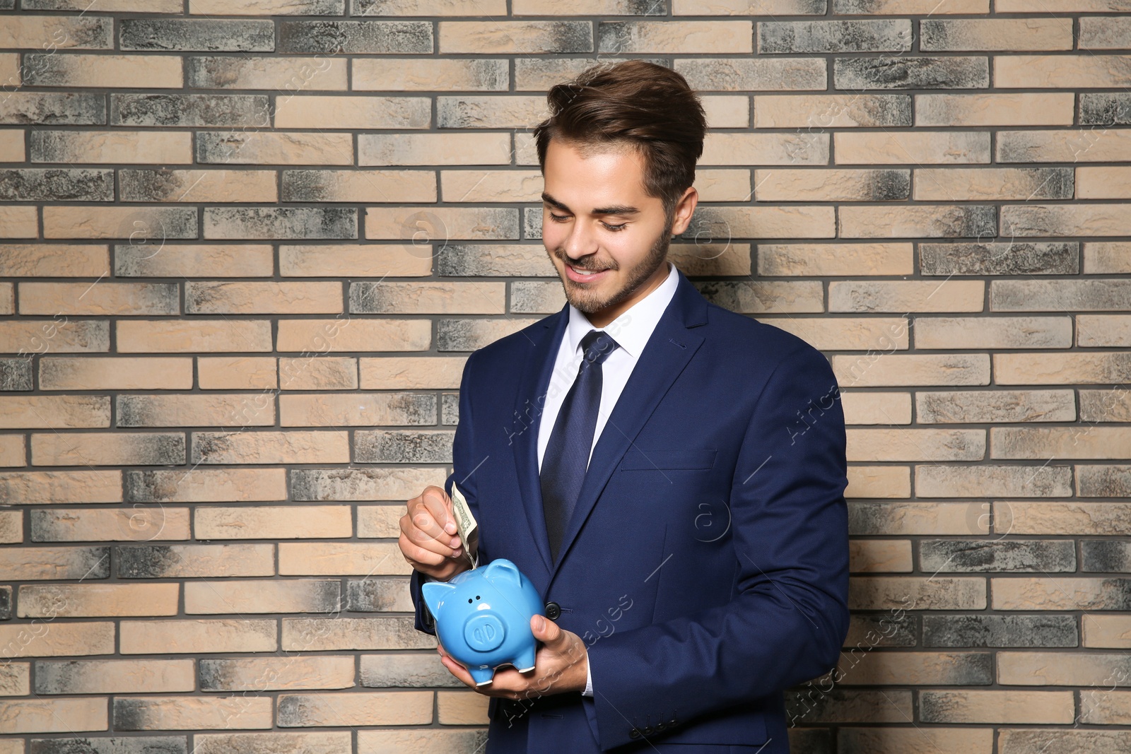 Photo of Young businessman putting money into piggy bank near brick wall