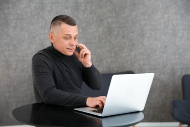 Photo of Mature man talking on mobile phone at table with laptop indoors