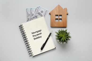 Photo of Notebook with words Mortgage Payment Holiday, house model, plant and money on light grey background, flat lay