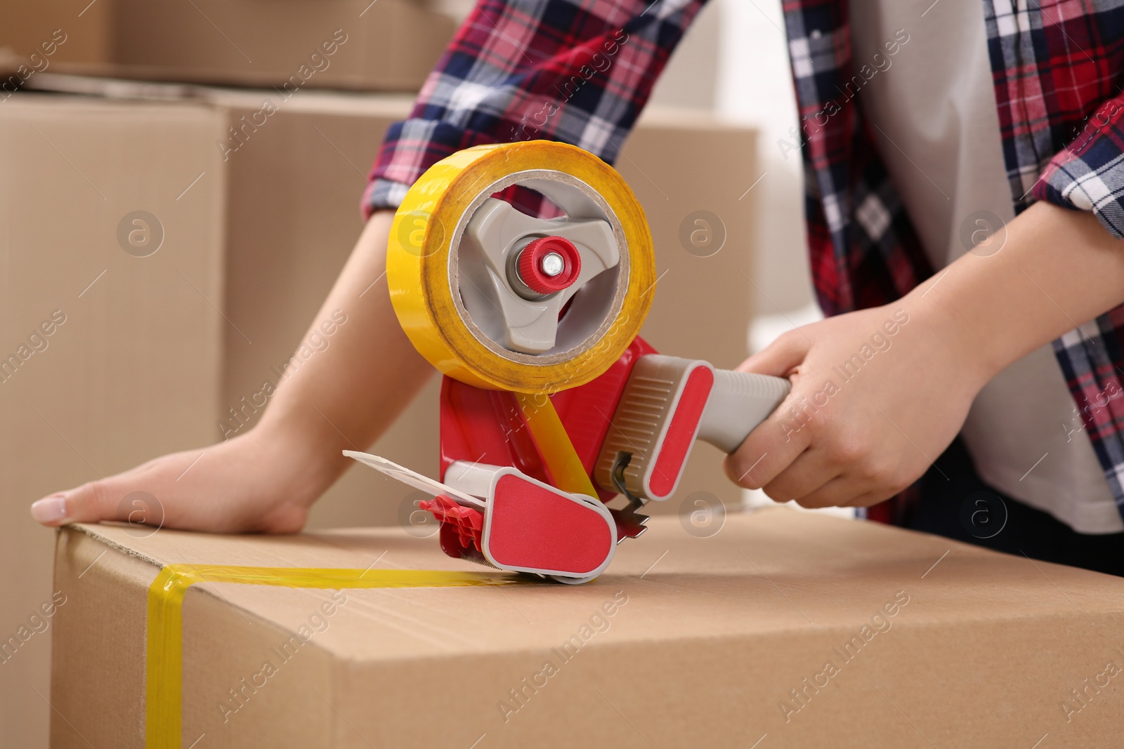 Photo of Woman applying adhesive tape on box with dispenser indoors, closeup