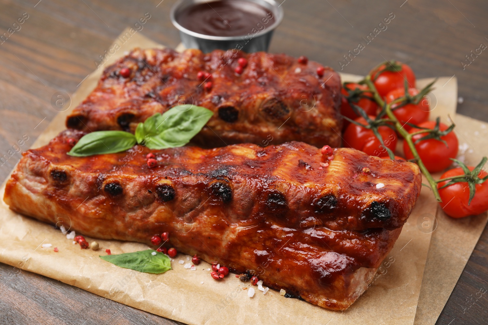 Photo of Tasty roasted pork ribs served with sauce, basil and tomatoes on wooden table, closeup