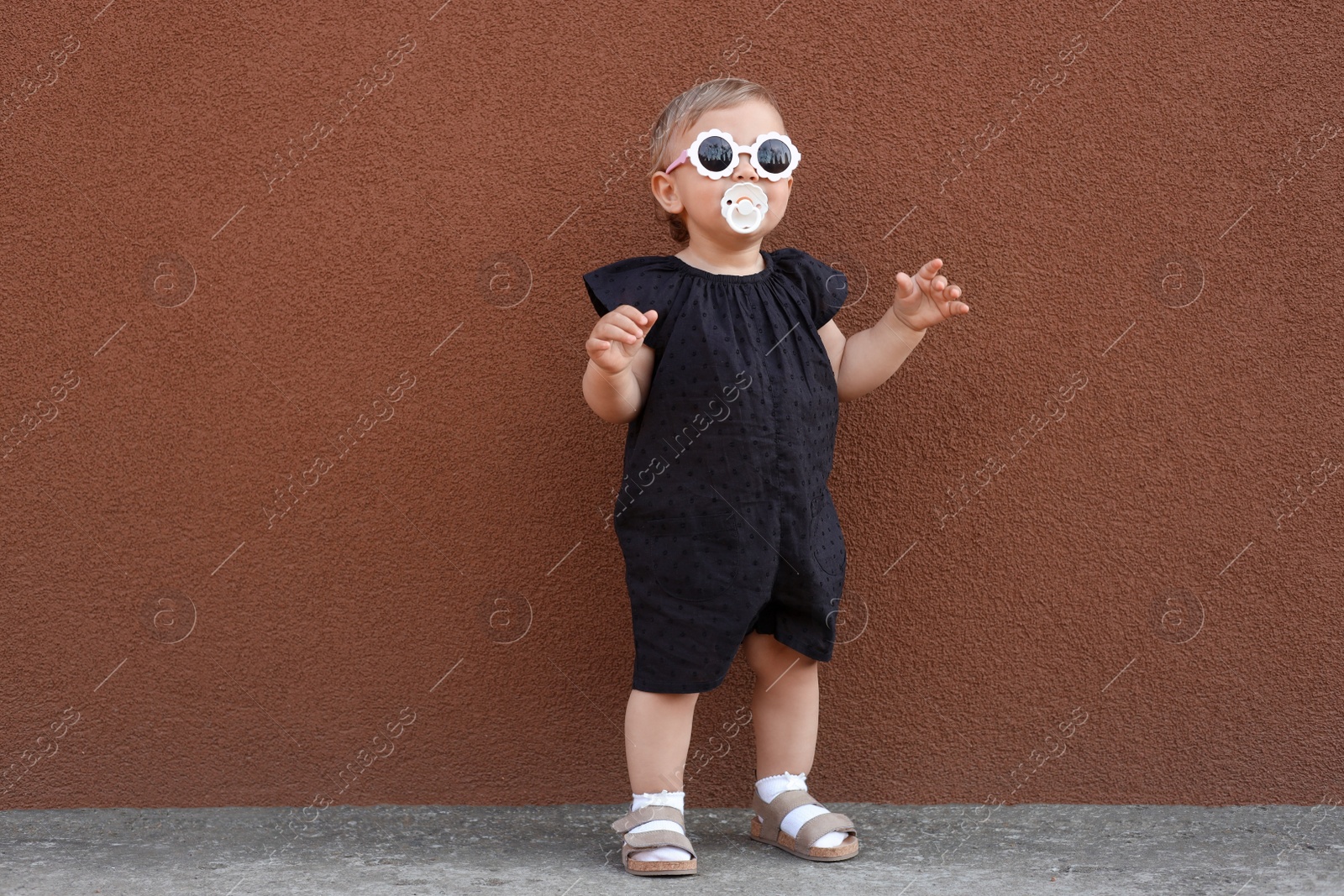 Photo of Cute little girl wearing stylish clothes with sunglasses and pacifier near brown wall outdoors