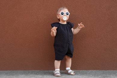Photo of Cute little girl wearing stylish clothes with sunglasses and pacifier near brown wall outdoors