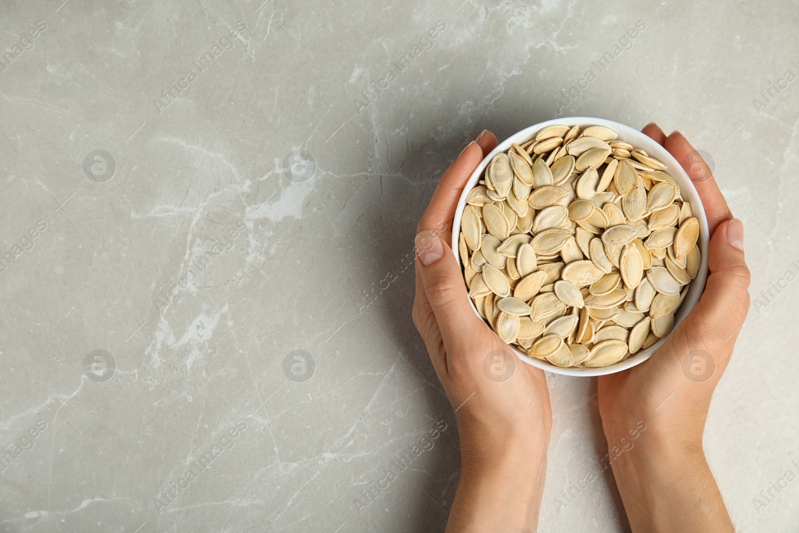 Photo of Young woman with bowl of raw pumpkin seeds at light grey marble table, top view. Space for text