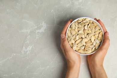 Photo of Young woman with bowl of raw pumpkin seeds at light grey marble table, top view. Space for text