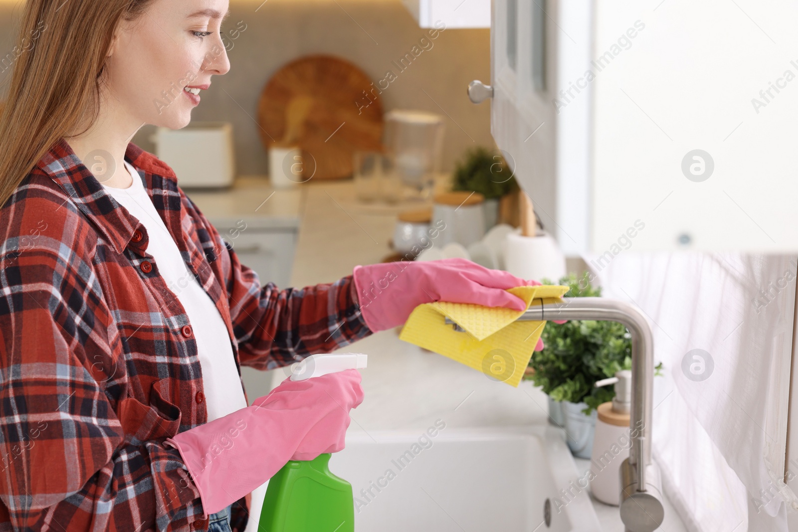 Photo of Woman with spray bottle and microfiber cloth cleaning water tap in kitchen