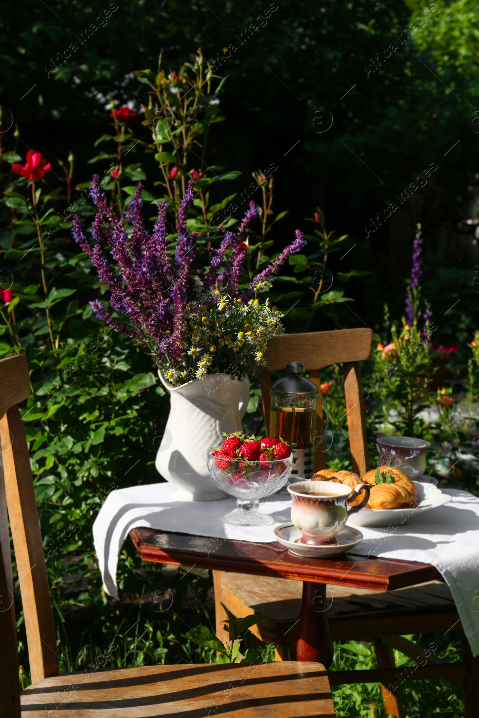 Photo of Beautiful bouquet of wildflowers, croissants and ripe strawberries on table served for tea drinking in garden