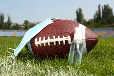 Photo of American football ball with hand sanitizer and protective mask on green field grass in stadium