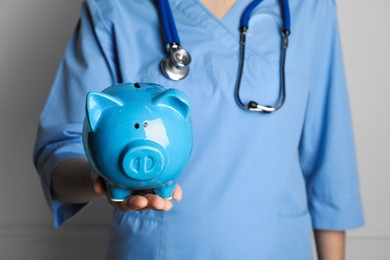 Doctor holding light blue ceramic piggy bank against white wall, closeup. Medical insurance