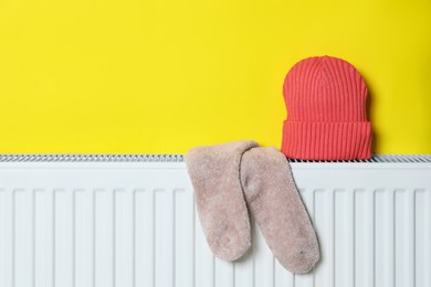 Knitted hat and socks on heating radiator near yellow wall, space for text